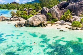 Vue aérienne d'une côte des Seychelles : blocs de granit, cocotiers et lagon turquoise, avec un kayak transparent