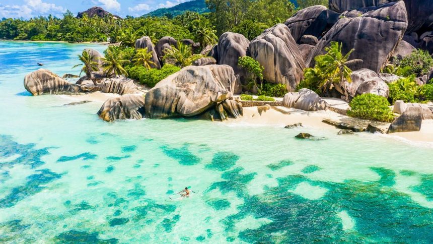 Vue aérienne d'une côte des Seychelles : blocs de granit, cocotiers et lagon turquoise, avec un kayak transparent